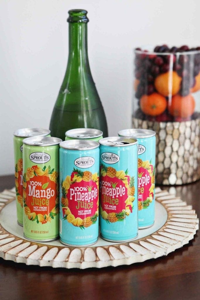 Sprouts Canned Mango and Pineapple Juices are displayed with champagne and a small fruit display on a table at an at-home holiday brunch