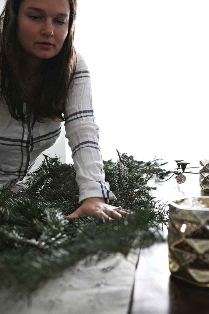 A woman decorates a holiday brunch table with fresh evergreen branches