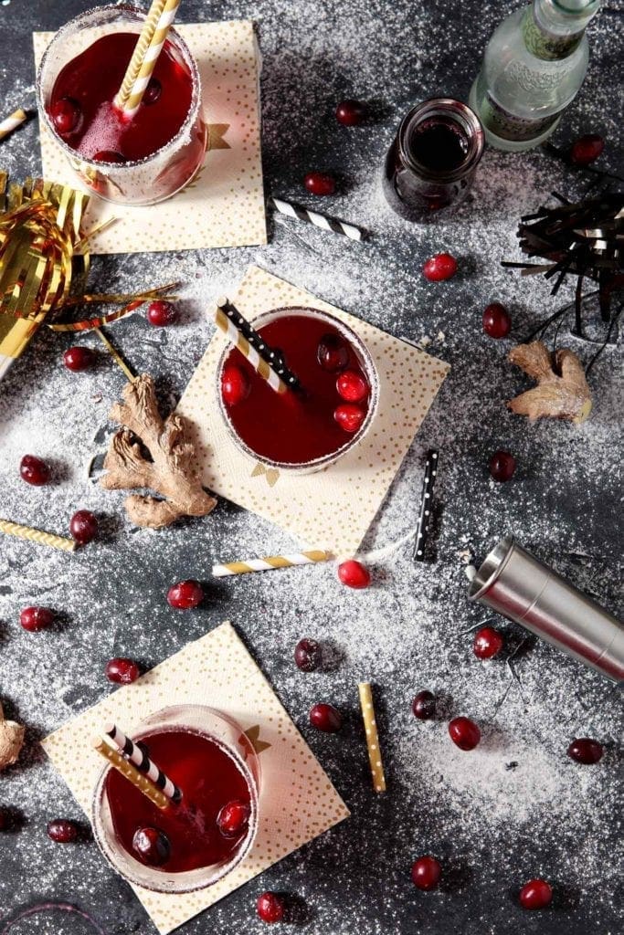 Three glasses of cranberry drinks are shown from above on a black board, surrounded by cranberries, ginger and "snow"