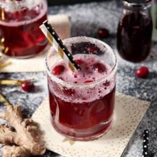 Two Sparkling Ginger Cranberry Mocktails are shown on a dark background, surrounded by fresh cranberries and ginger