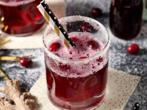 Two Sparkling Ginger Cranberry Mocktails are shown on a dark background, surrounded by fresh cranberries and ginger