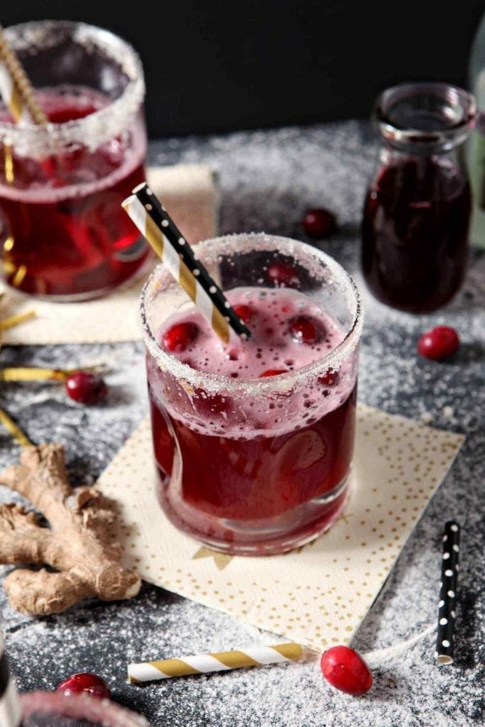 Two Sparkling Ginger Cranberry Mocktails are shown on a dark background, surrounded by fresh cranberries and ginger