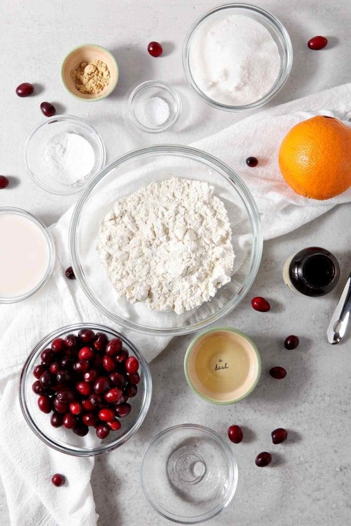 Ingredients for Vegan Orange Cranberry Muffins are shown on a grey tabletop, surrounded by fresh cranberries