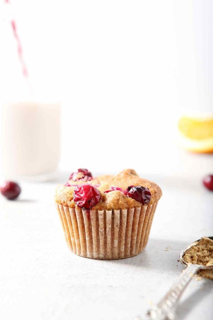 One Vegan Orange Cranberry Muffin sits in front of a glass of non-dairy milk, surrounded by fresh cranberries and orange slices