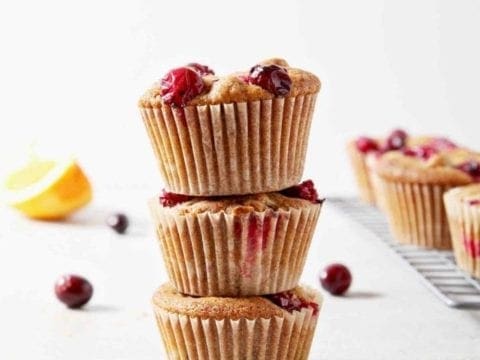A stack of three Vegan Orange Cranberry Muffins stand beside a wire cooling rack with more muffins