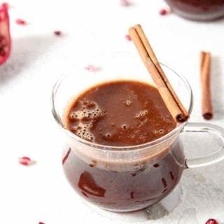 A close up of a glass mug holding Pomegranate Hot Buttered Rum, garnished with a cinnamon stick