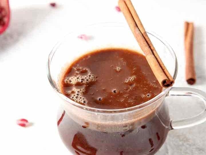 A close up of a glass mug holding Pomegranate Hot Buttered Rum, garnished with a cinnamon stick