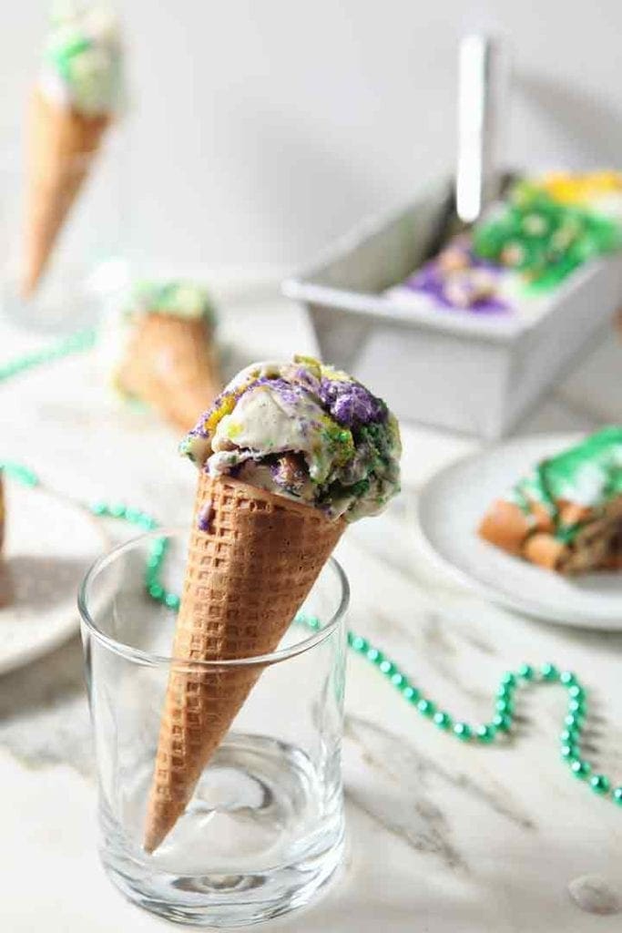 A cone of No Churn Mardi Gras King Cake Ice Cream, with other cones, ice cream and king cake slices in the background