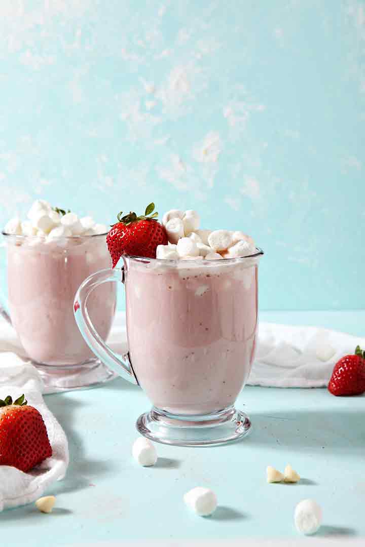 Two mugs of Strawberry White Hot Chocolate are garnished with marshmallows and strawberries