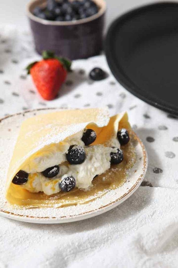 A blueberry crepe is served on a plate, sprinkled with powdered sugar and fresh whipped cream