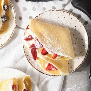 Homemade breakfast crepes are shown from above, sprinkled with powdered sugar and filled with whipped cream and fruit, with a Swiss Diamond crepe pan