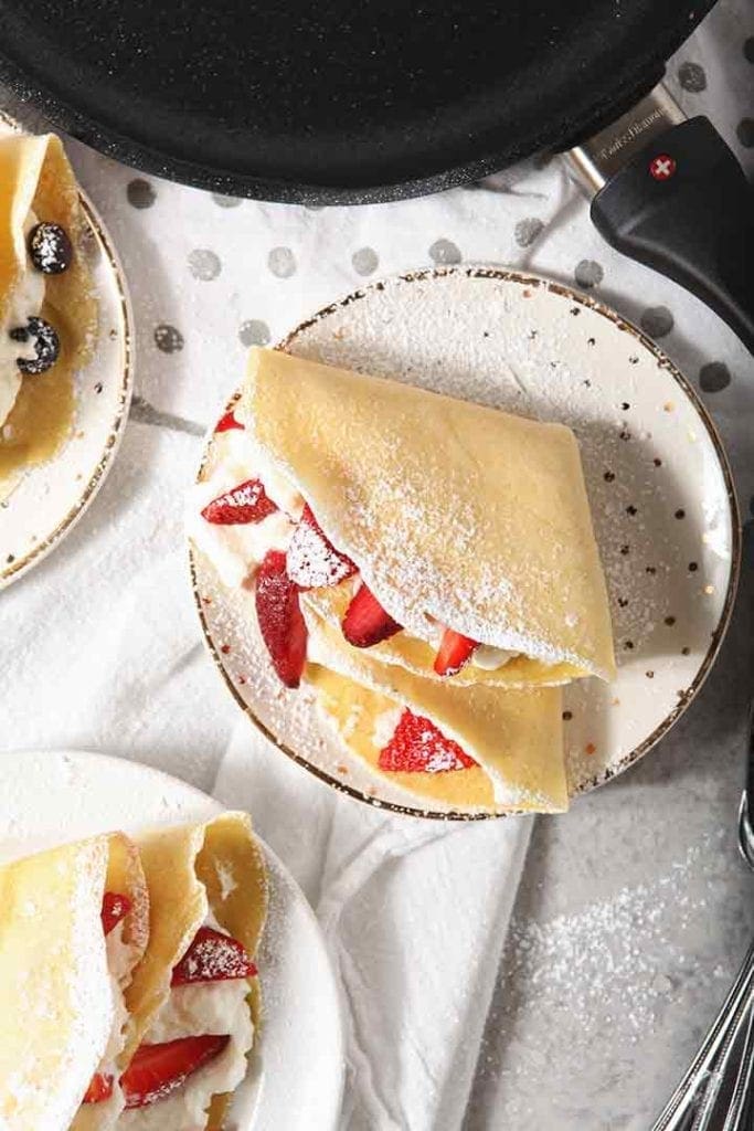 Homemade breakfast crepes are shown from above, sprinkled with powdered sugar and filled with whipped cream and fruit, with a Swiss Diamond crepe pan