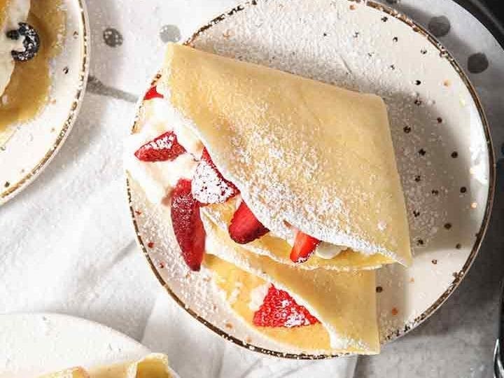 Homemade breakfast crepes are shown from above, sprinkled with powdered sugar and filled with whipped cream and fruit, with a Swiss Diamond crepe pan