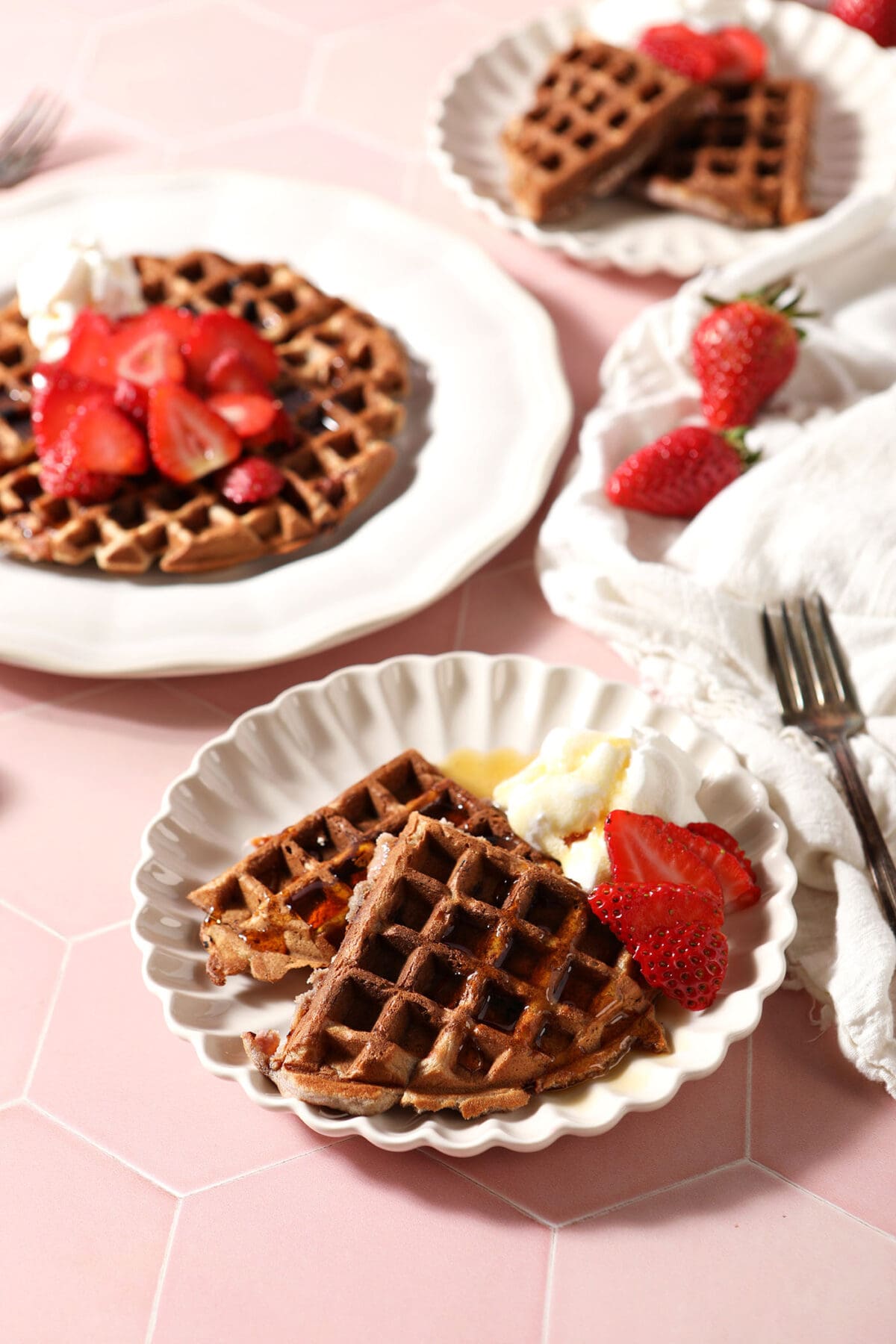 Three plates of strawberry waffles on a pink tiled surface with a white linen and fresh strawberries