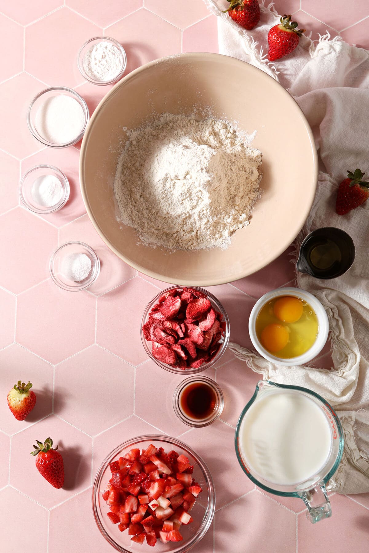 Bowls of ingredients to make waffles with fresh and freeze-dried strawberries on a pink tiled surface next to a white linen