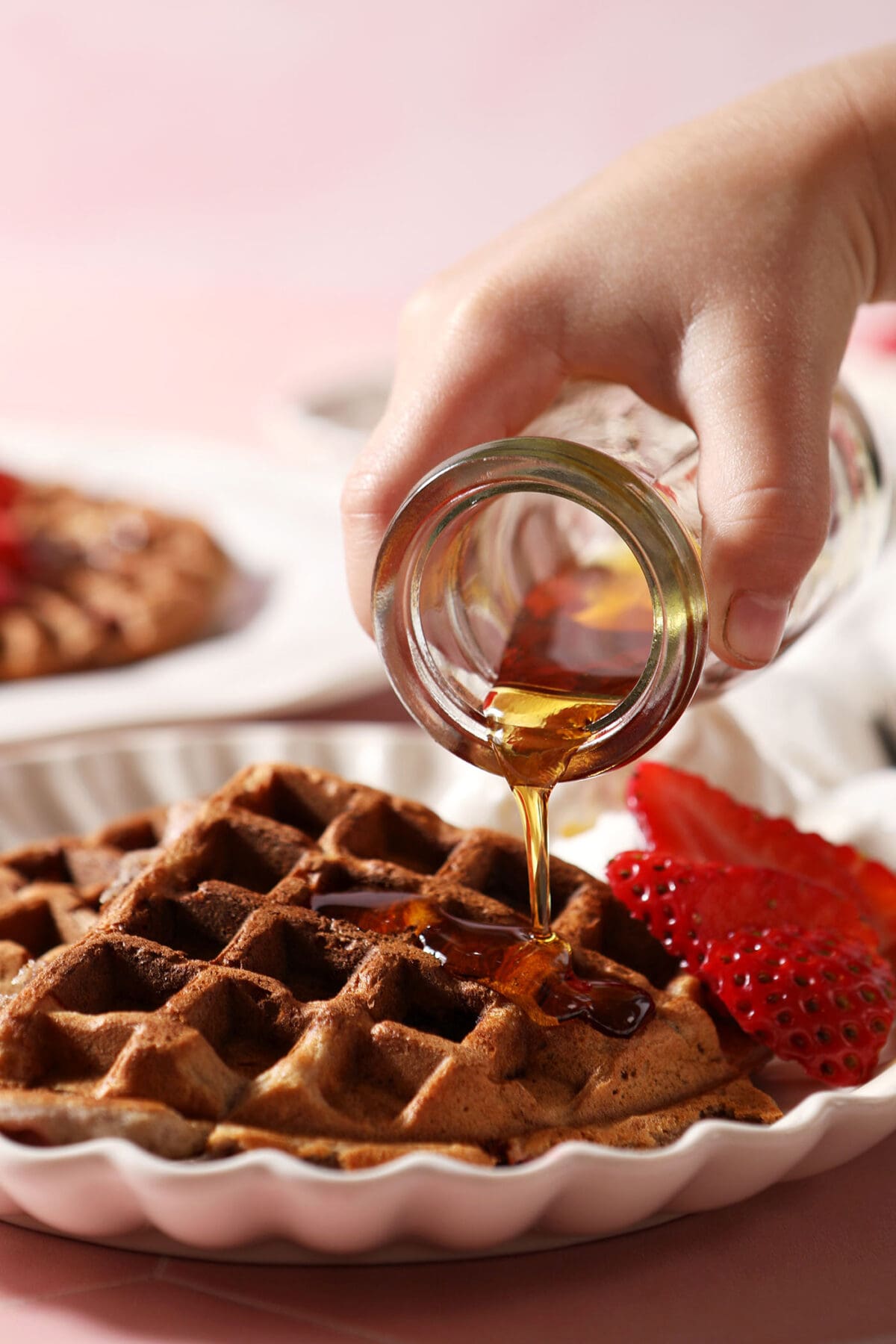 A child pours maple syrup onto a waffle next to sliced strawberries