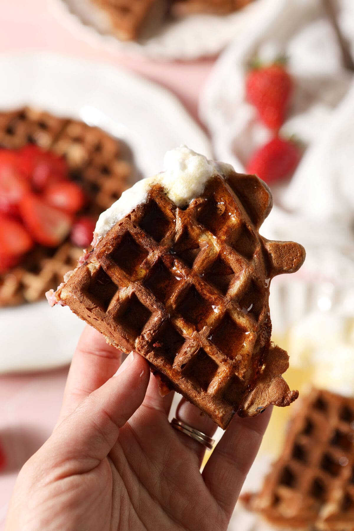 A hand holds a portion of a waffle dipped in whipped cream above plates of more waffles on a pink surface