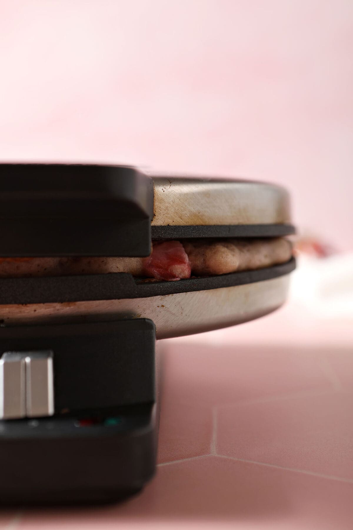 Waffle batter cooks in a waffle iron on a pink tiled surface