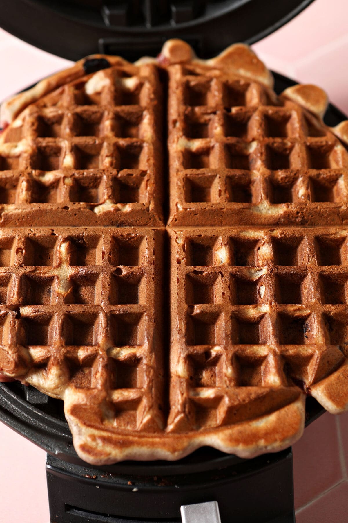 A cooked strawberry waffle in a waffle iron on a pink surface