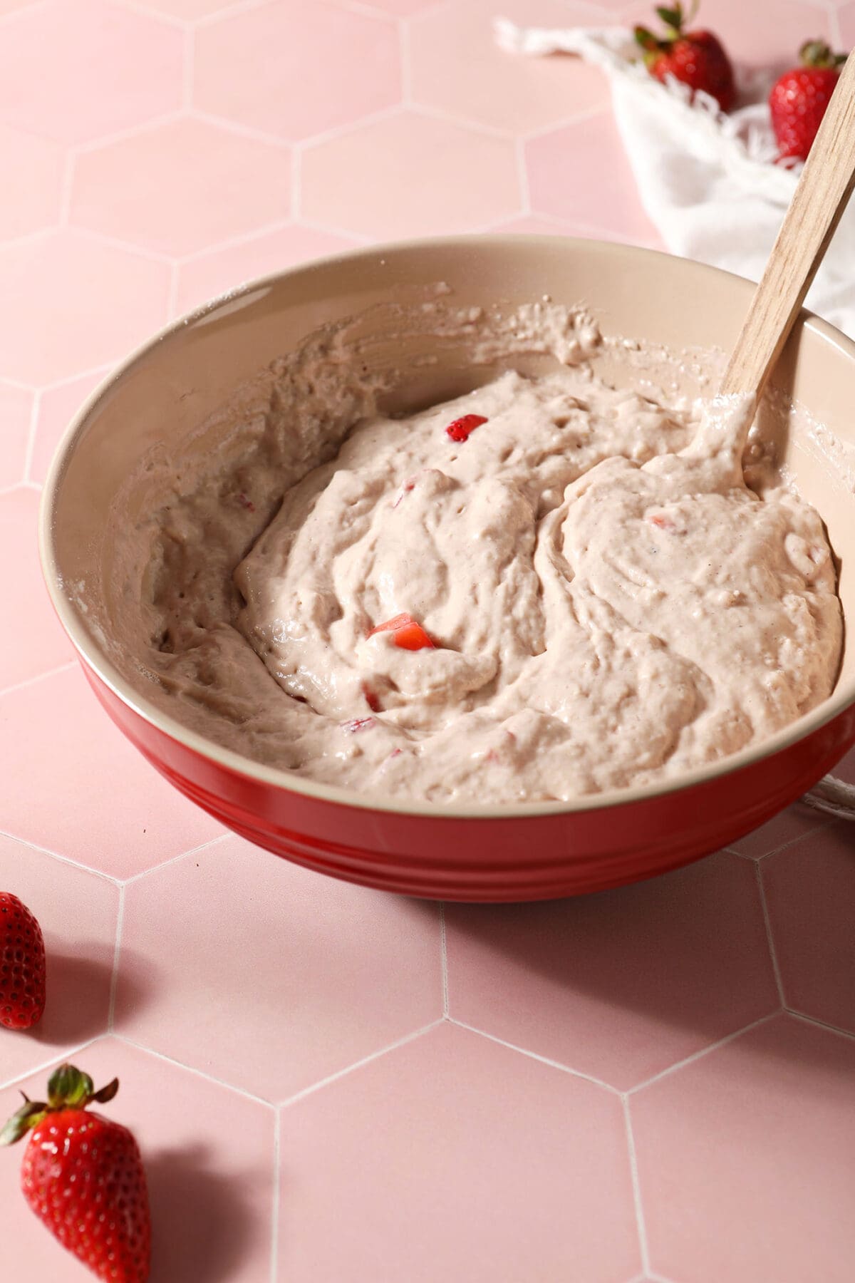 Mixed strawberry waffle batter in a red bowl on a pink tiled surface surrounded by fresh strawberries and a white linen