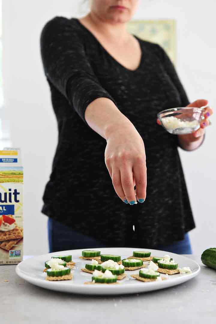 A woman sprinkles feta cheese over a cucumber appetizer