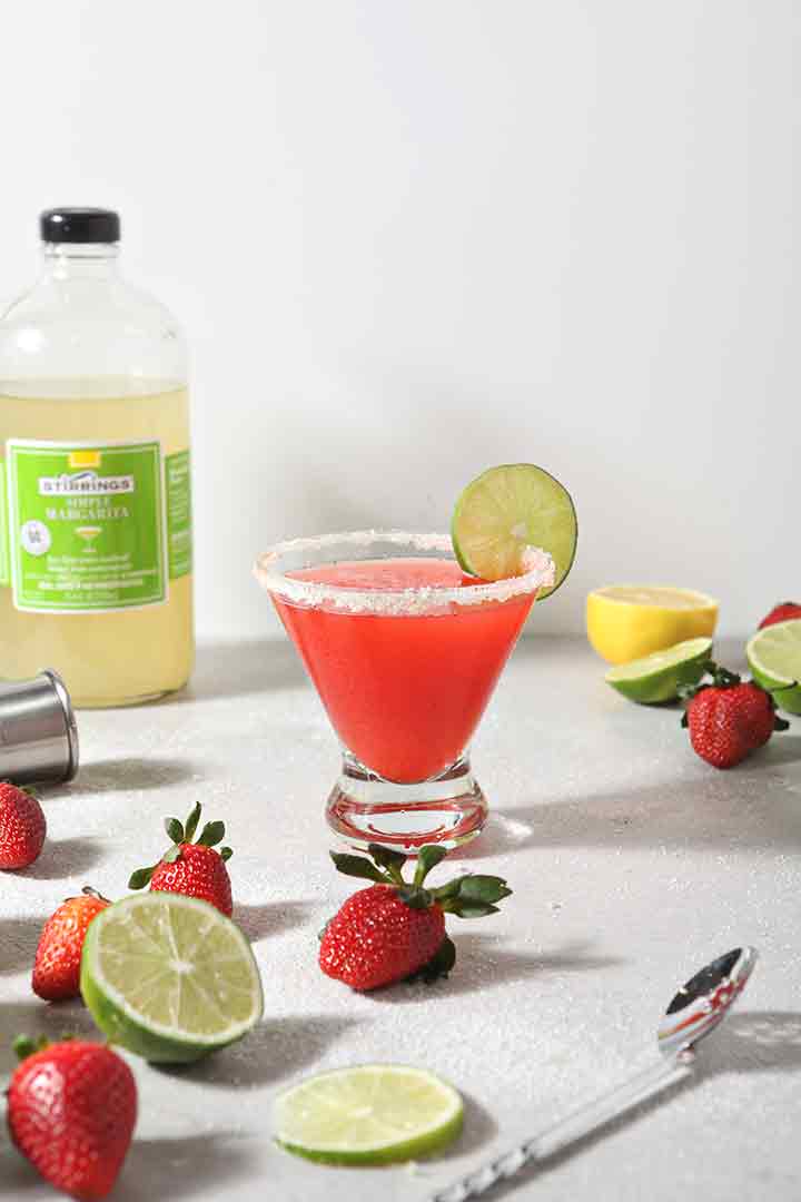 A pink margarita sits surrounded by fresh strawberries and limes