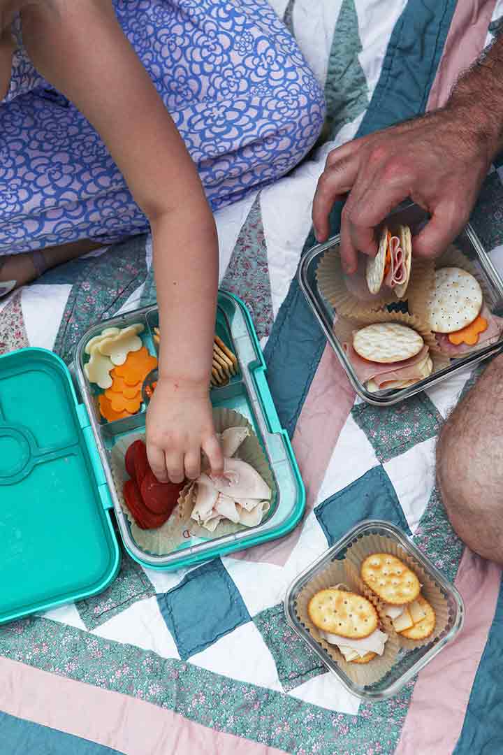 A girl and a man eat cracker sandwiches out of portable storage containers on a picnic