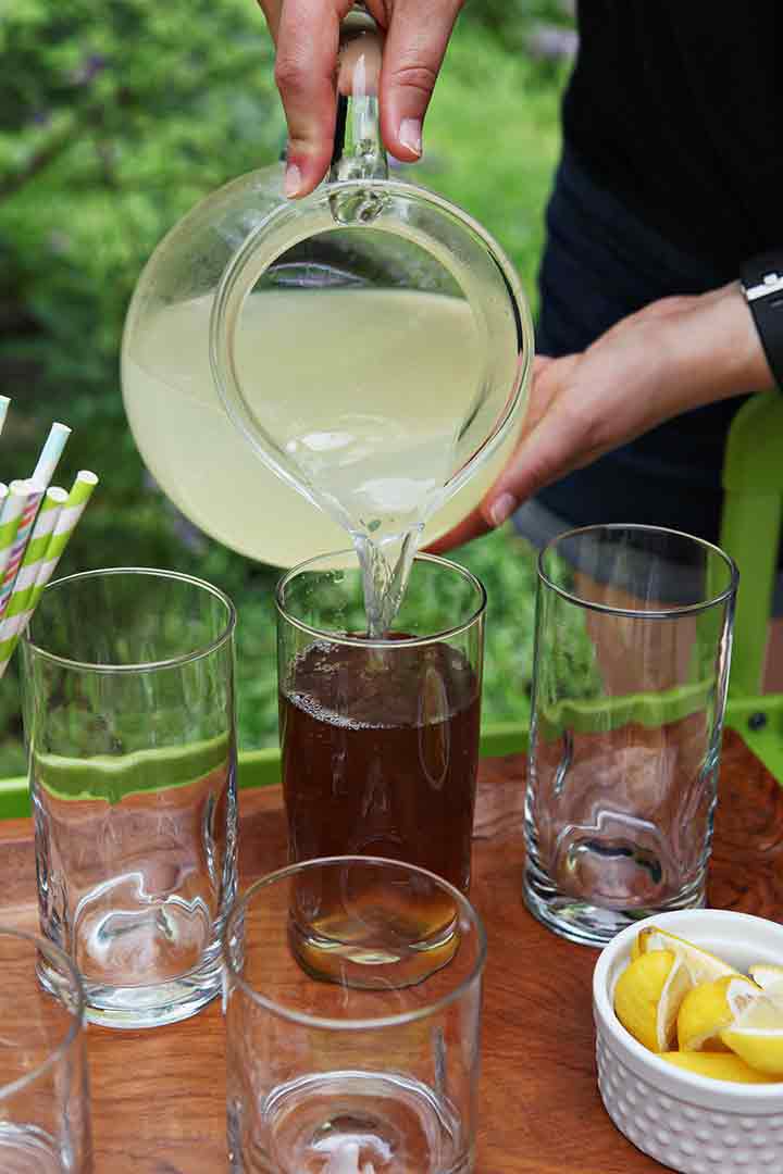 A woman pours lemonade on top of tea in a glass while entertaining outdoors