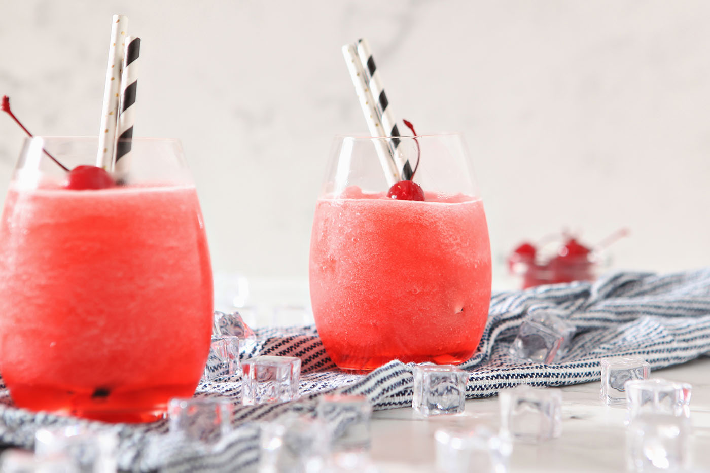 Two glasses of cherry bourbon slush garnished with maraschino cherries and straws sit on a blue and white striped linen surrounded by ice on a marble surface