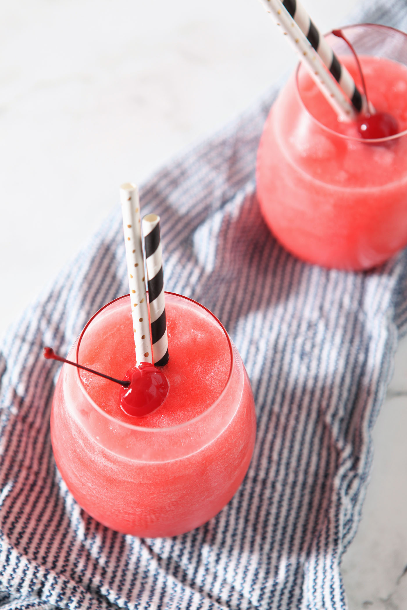 Overhead of two bourbon cherry slushes sitting on top of a blue and white striped linen