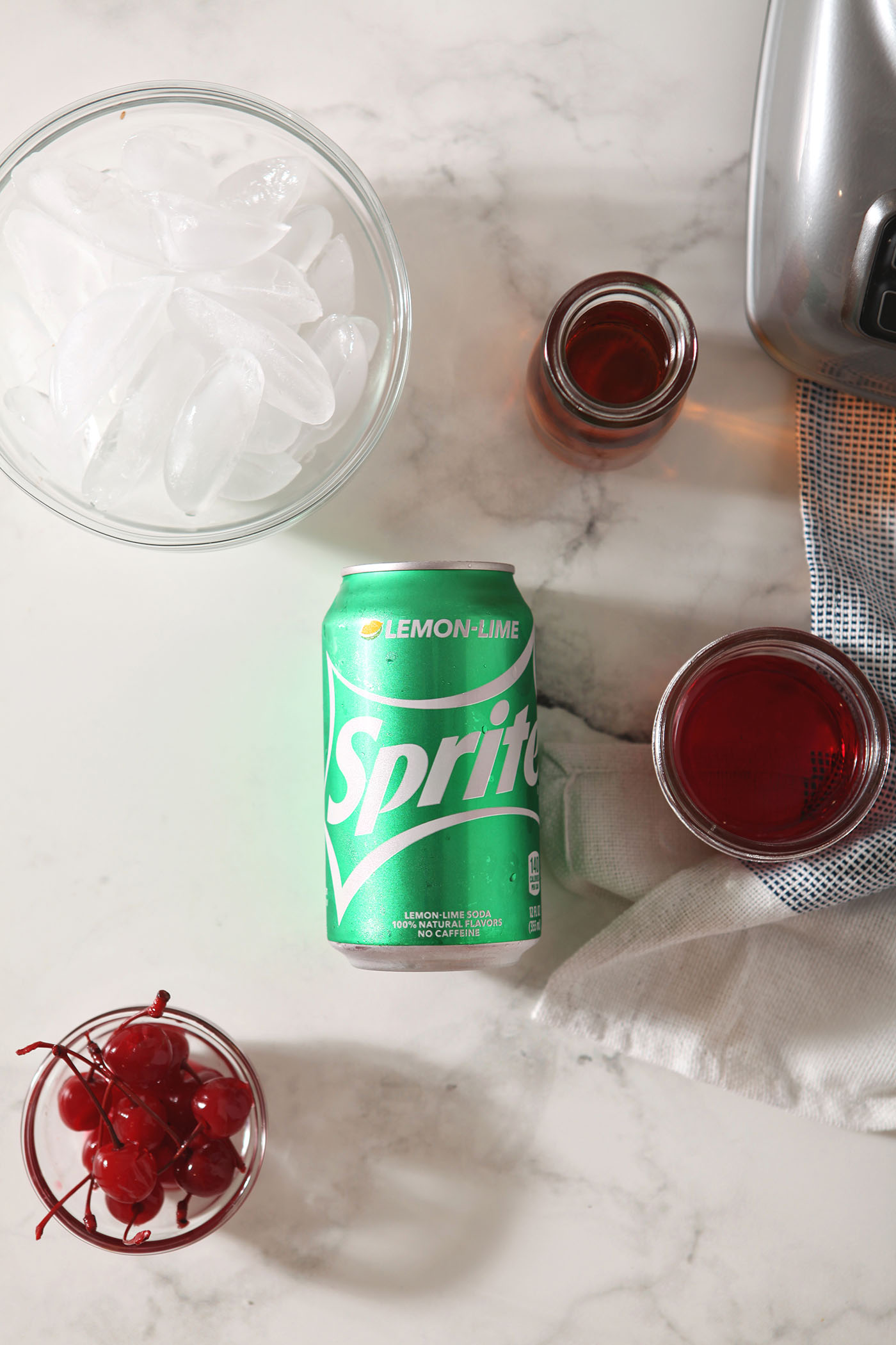 A bowl of cherries, cherry juice, a sprite, bourbon and ice in containers on a marble surface to make slushes