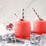 Two wine glasses holding bourbon cherry slushes garnished with maraschino cherries and straws, sit on a blue and white striped linen, surrounded by ice cubes on a marble surface