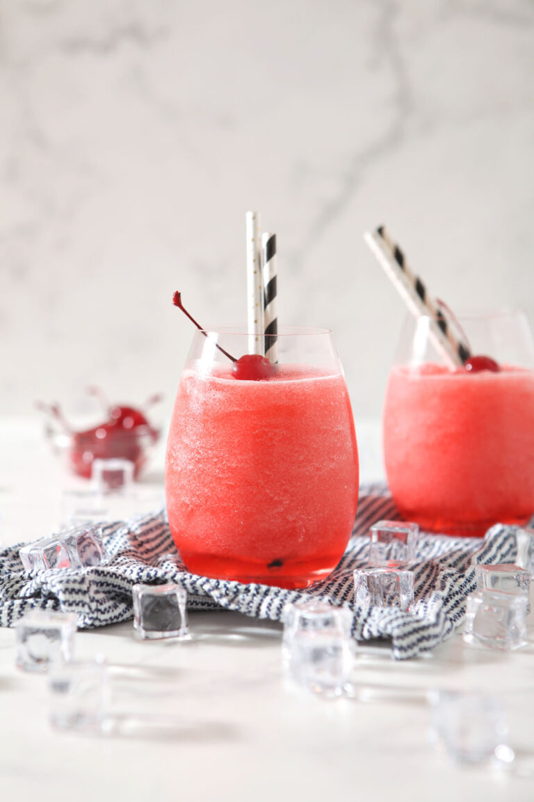 Two wine glasses holding bourbon cherry slushes garnished with maraschino cherries and straws, sit on a blue and white striped linen, surrounded by ice cubes on a marble surface