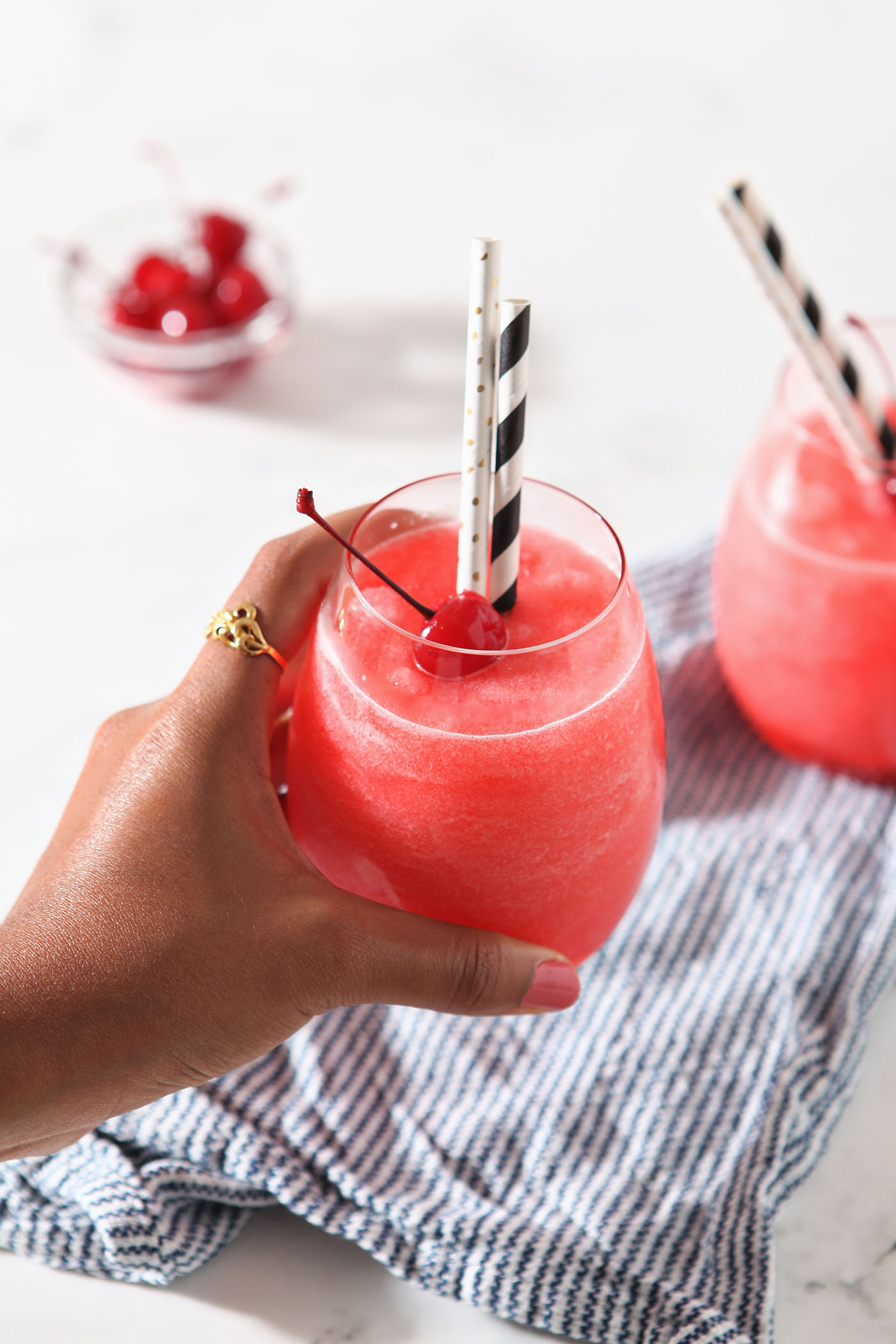 A hand holds a cherry slush above a marble surface with a blue and white striped linen and another glass of slush