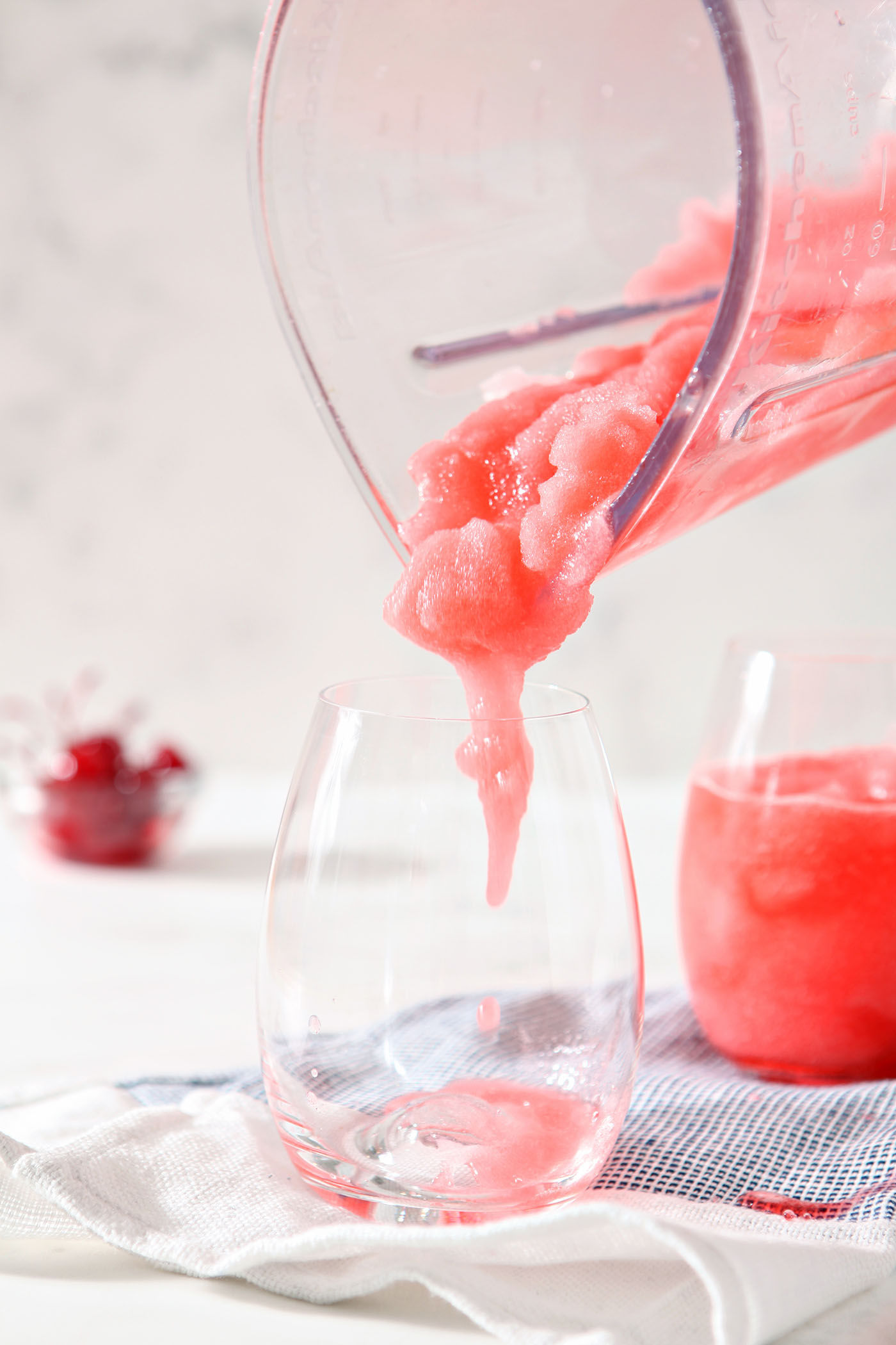 A slush with cherries and bourbon pours into a wine glass on a blue and white striped linen