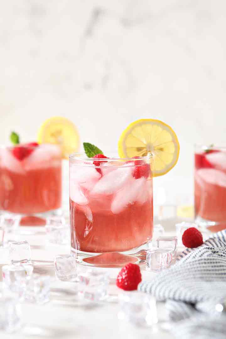 Three glasses of Spiked Raspberry Lemonade are surrounded by ice, raspberries and lemons