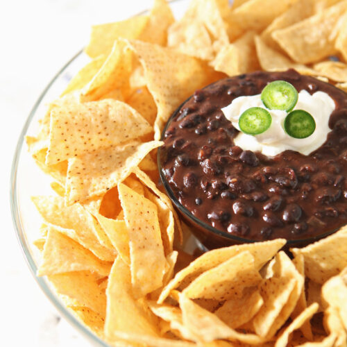 Chips surround a bowl of black bean dip, garnished with sour cream and jalapeno slices.