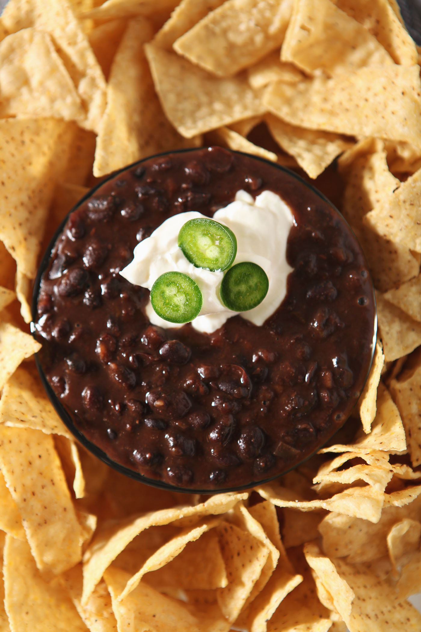 Close-up of a bowl of black bean dip garnished with sour cream and jalapeno slices and surrounded by tortilla chips