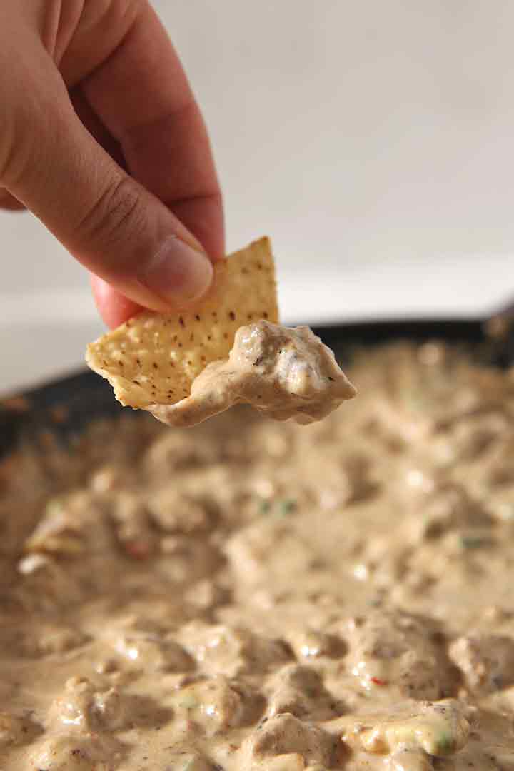 A close up of a woman lifting dip up out of the skillet with a tortilla chip