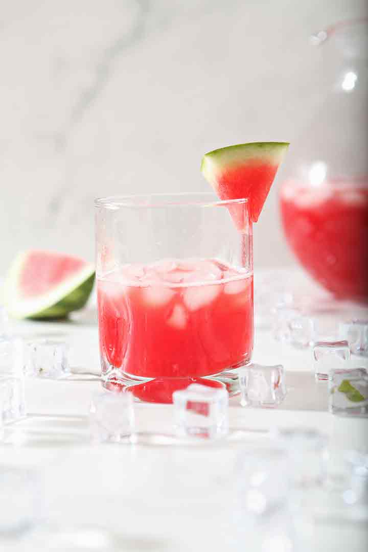 A singular glass is shown, garnished with a watermelon wedge and surrounded by ice