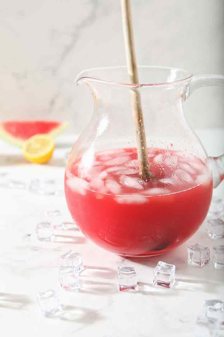 A wooden spoon stirs ice cubes and juice around a pitcher