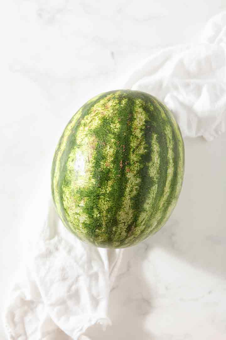 A whole watermelon sits on a marble surface, surrounded by a white tea towel