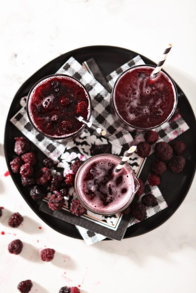 Three purple potion drinks on a black plate with blackberries