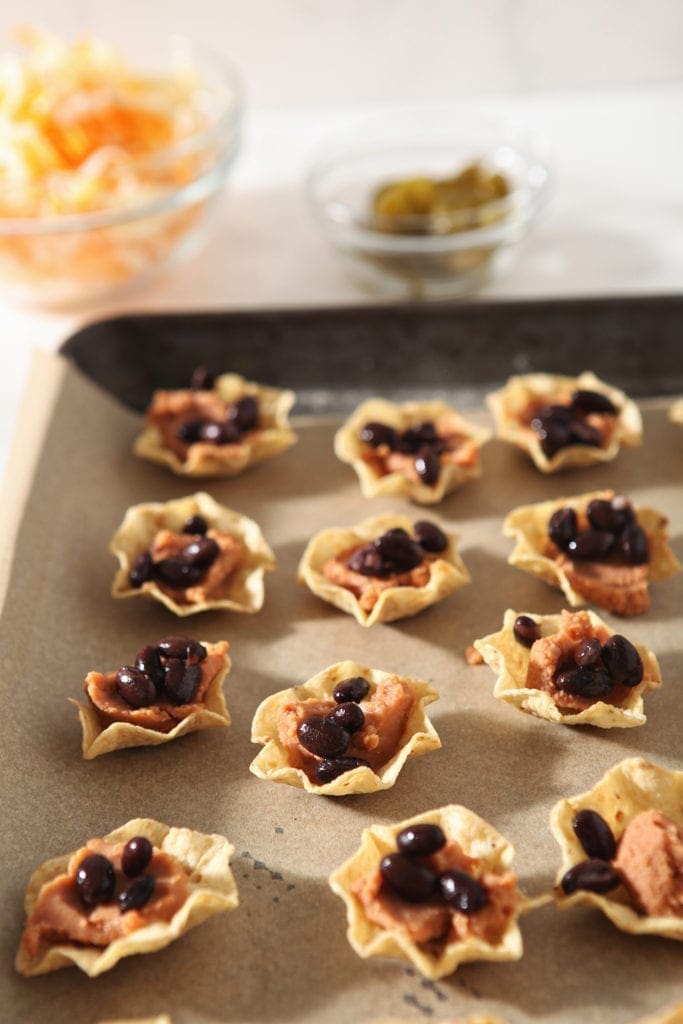 Tortilla chips on a baking sheet are filled with refried beans and black beans to make individual nachos