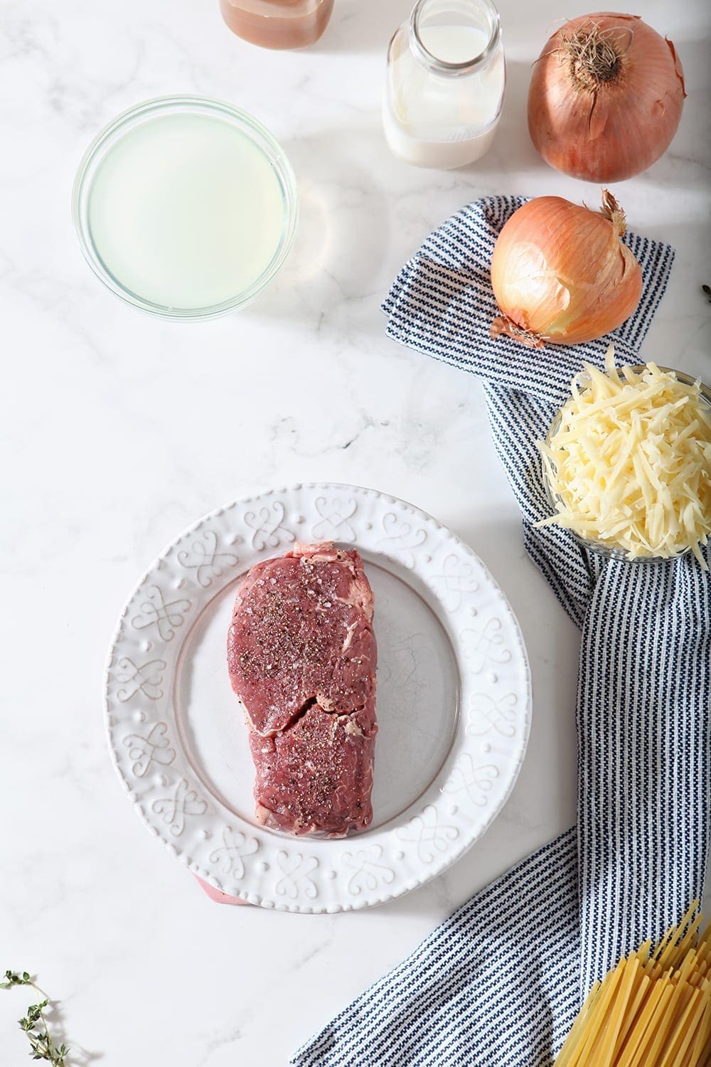 A Panorama ribeye, seasoned with salt and pepper, sits on a white plate, with other ingredients, before cooking