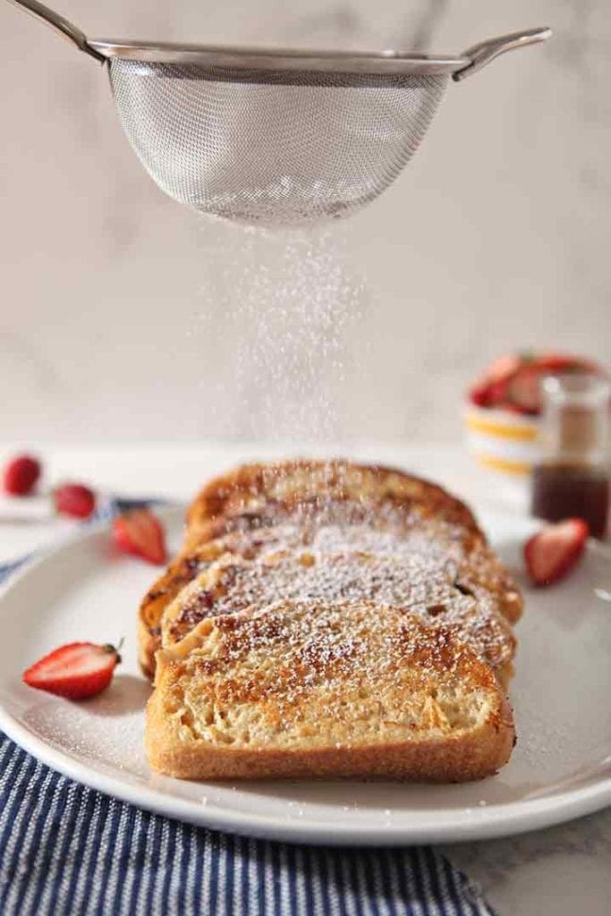 Powdered sugar is sprinkled on top of the French toast on a white serving platter