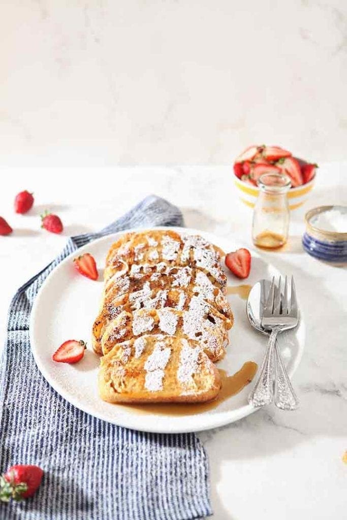 Yogurt French Toast, on a white plate, drizzled with maple syrup and sprinkled with powdered sugar, with strawberries