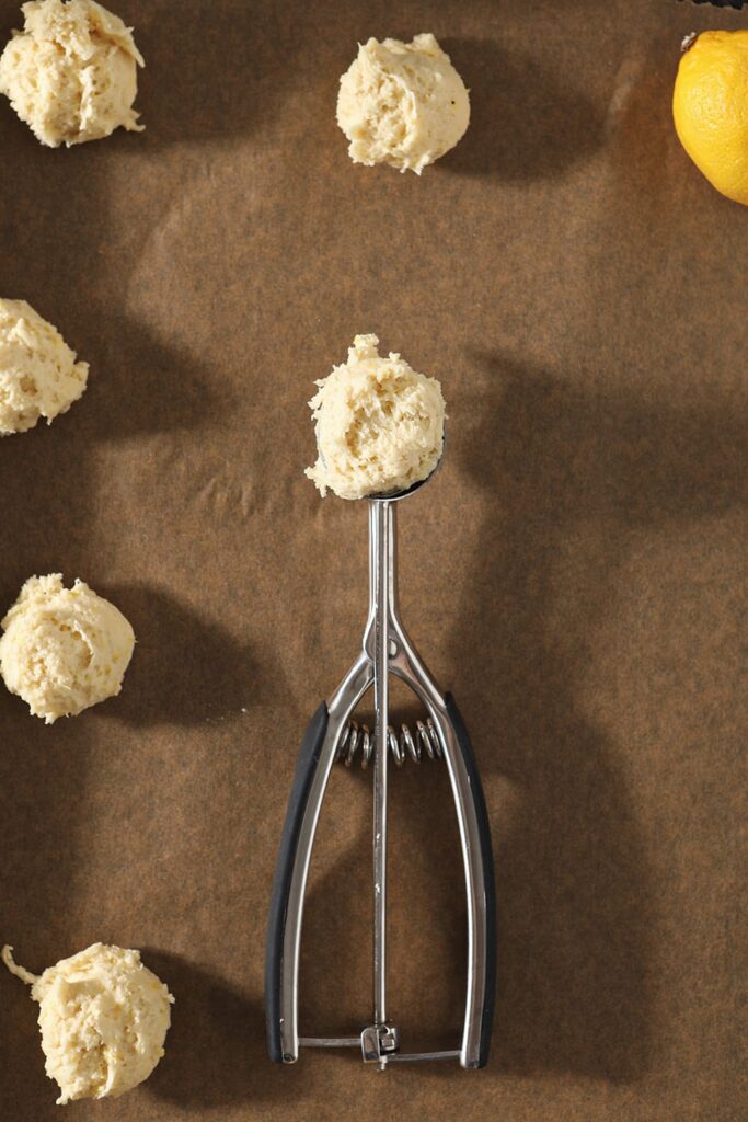 A cookie scoop holds a portion of lemon drop cookie batter on a baking sheet next to scooped cookies