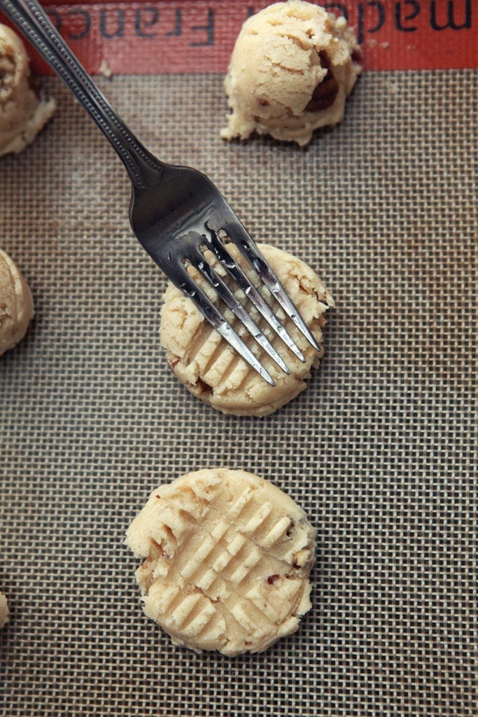 A fork's tines press into a ball of cookie dough on a baking sheet