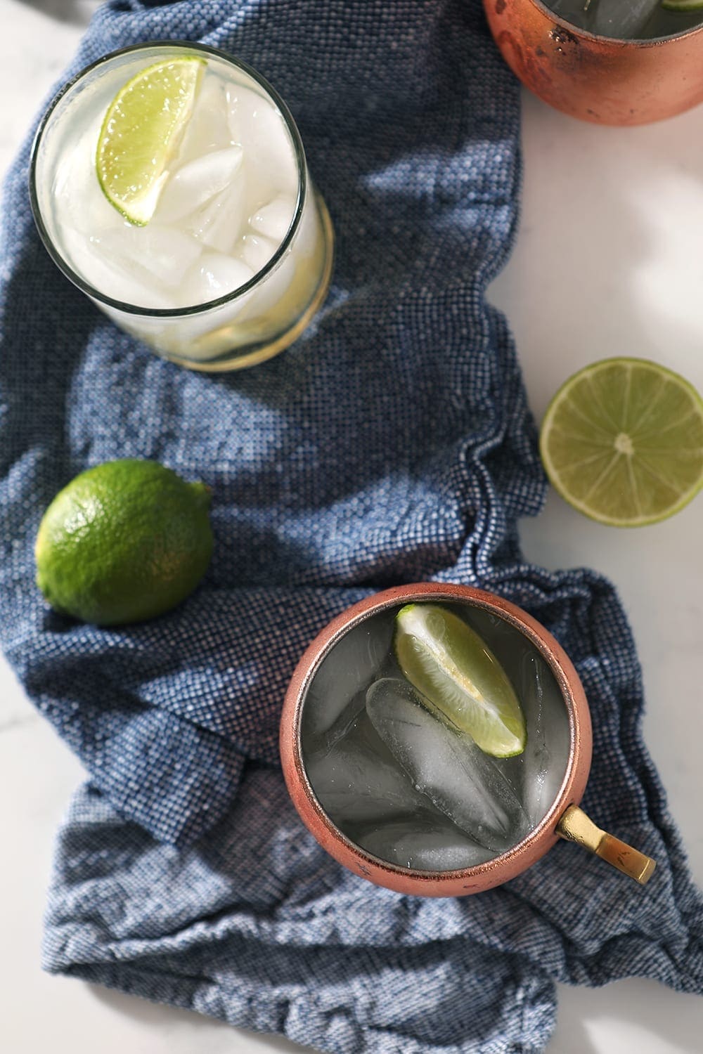 Overhead of three drinks on a blue towel, with limes scattered around them